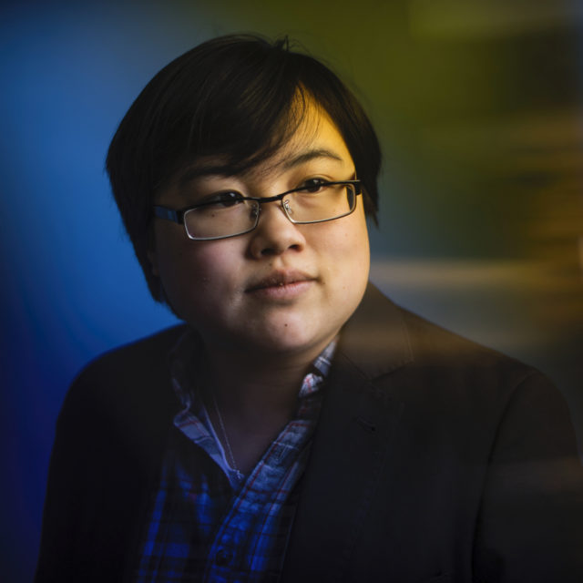 Headshot image of Lydia X. Z. Brown, an east-asian, non-binary individual with short black hair, glasses, a blue plaid shirt and black blazer.
