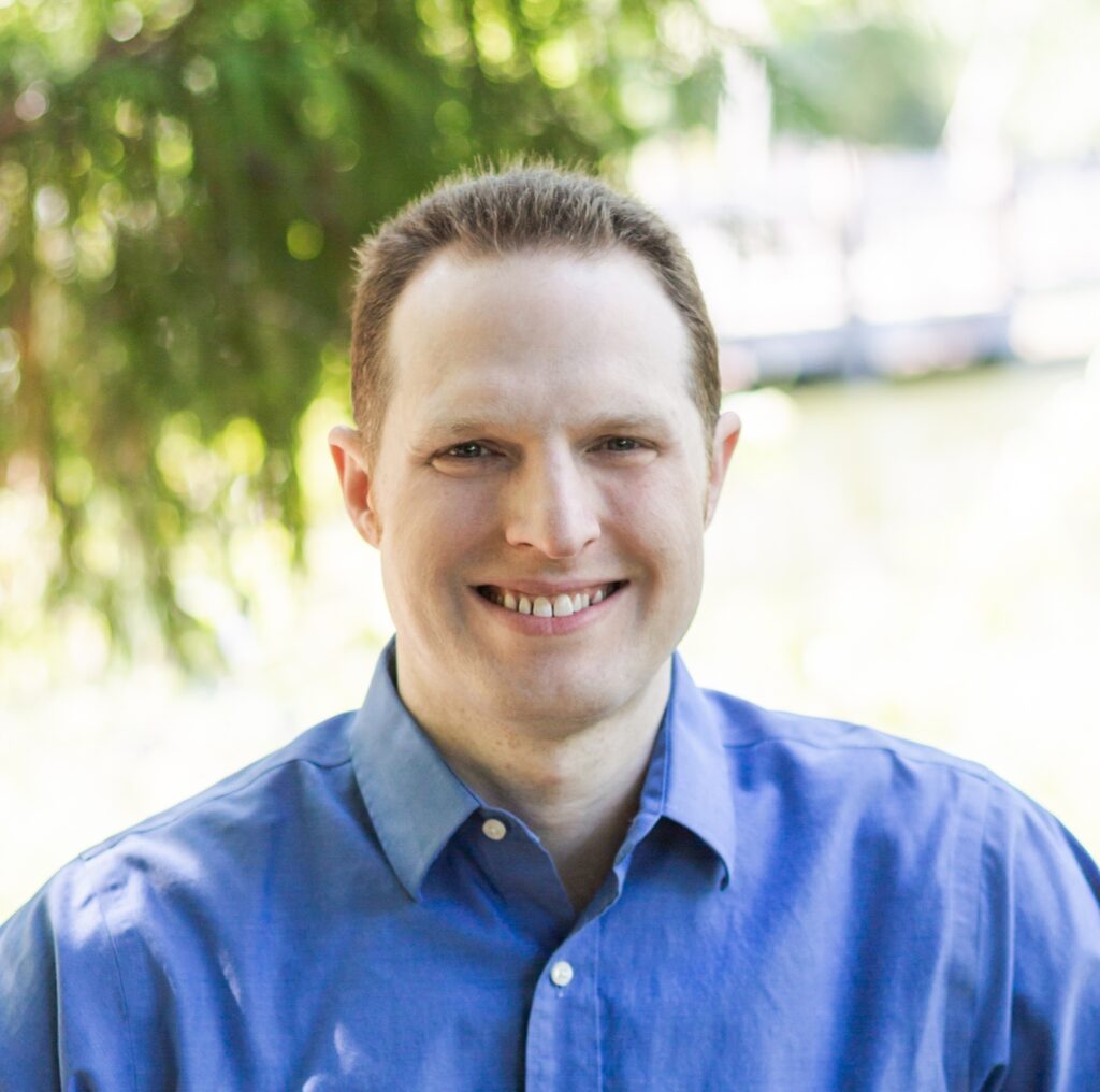 Matt Scherer, smiling in a blue collared shirt, outside in front of a green tree.