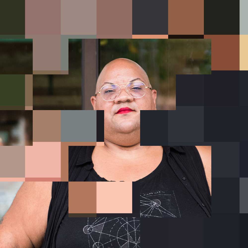 Original photo by Chona Kasinger for the Disabled And Here project. A black non-binary person stands casually outside a cafe, dressed in all black with a shaved head, glasses, a red lip, along with moon earrings. https://affecttheverb.com/disabledandhere/