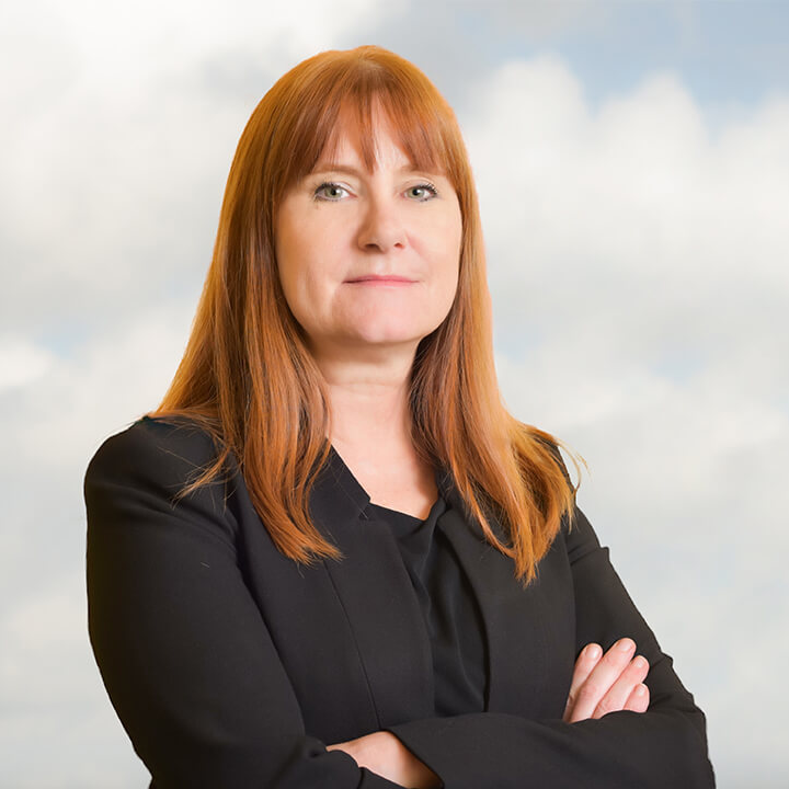 Image of Eve Hill, with red hair and wearing a black suit with arms crossed. In front of a cloudy sky background.