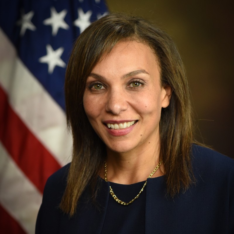 Image of Rebecca Bond, wearing a dark top and necklace in front of an American flag and dark background.