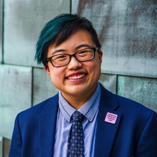 Photo: Ly Xīnzhèn M. Zhǎngsūn Brown smiles and tilts their head slightly to the side, looking confidently at the camera. They are a young-ish East Asian person with a streak of teal in their short black hair, wearing glasses, a cobalt blue jacket and navy tie, with a blue copper wall behind them. Photo by Sarah Tundermann.