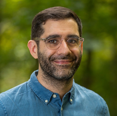 Solon Barocas. Assistant Professor, Cornell University. Wearing glasses and a blue collared shirt, in front of a green natural background.