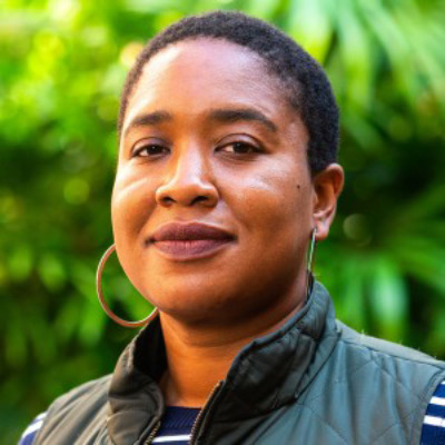Jasmine McNealy. Associate Professor, University of Florida. Wearing hoop earrings and a green vest over a blue and white striped shirt. Green leafy background.