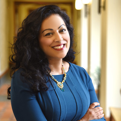 Shobita Parthasarathy. Professor and Director, Science, Technology, and Public Policy Program, University of Michigan. Wearing a blue dress and a gold necklace, and standing within a long hallway.