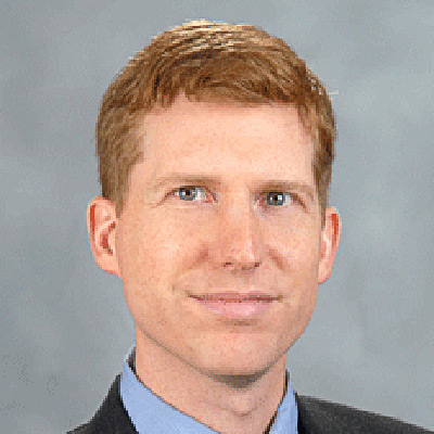 Robert Seamans. Associate Professor of Management and Organizations, New York University. Wearing a blue shirt and grey suit, in front of a light blue backdrop.