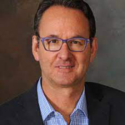 Henry Claypool. Independent. Wearing glasses and a light blue collared shirt with a dark suit, in front of a dark grey backdrop.