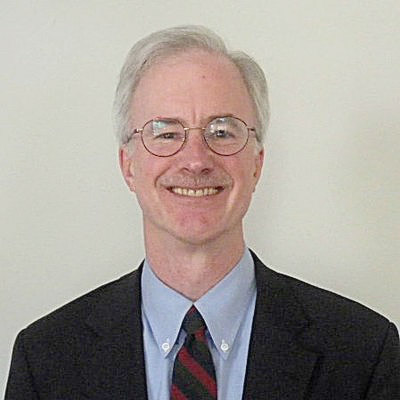 George Slover. Wearing glasses, a blue collared shirt and dark suit, in front of a light colored wall.