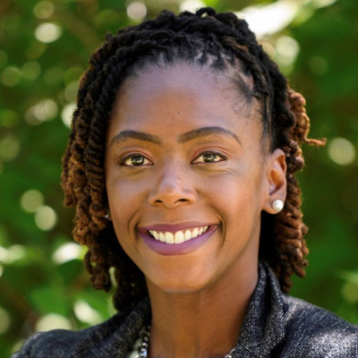 Dominique Harrison. Joint Center for Political and Economic Studies. Wearing a dark grey shirt, in front of a leafy green background.