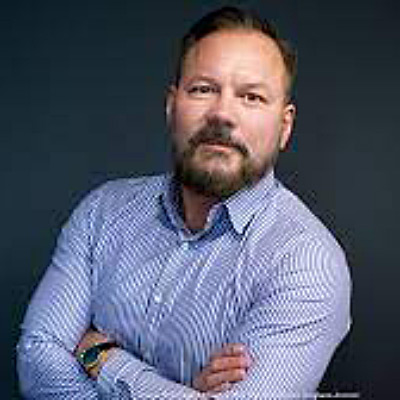 Brian Huseman. Amazon. Wearing a light blue collared shirt, in front of a dark blue backdrop.