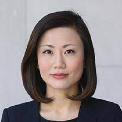 Denise Zheng. Wearing a dark grey suit, in front of a light grey background.