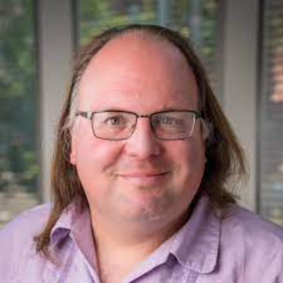Ethan Zuckerman. Massachusetts Institute of Technology. Wearing a bright purple collared shirt, in front of a leafy green background.
