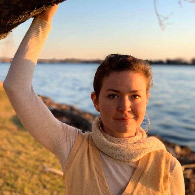 Yona Tali Roberts Golding. Wearing hoop earrings, a white scarf wrapped around their neck, and a white shirt with a light colored sweater vest over it. Fading orange sunlight from the right, with a bright blue sky and blue body of water behind them. Arm extended above, leaning against a tree.