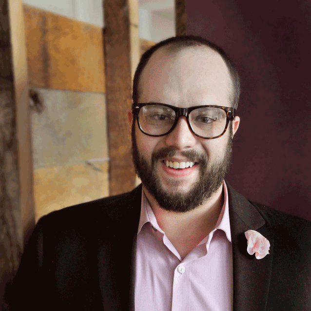 Jake Laperruque. Wearing a pink collared shirt and a pink flower lapel pin on a dark suit jacket, with dark rimmed glasses.