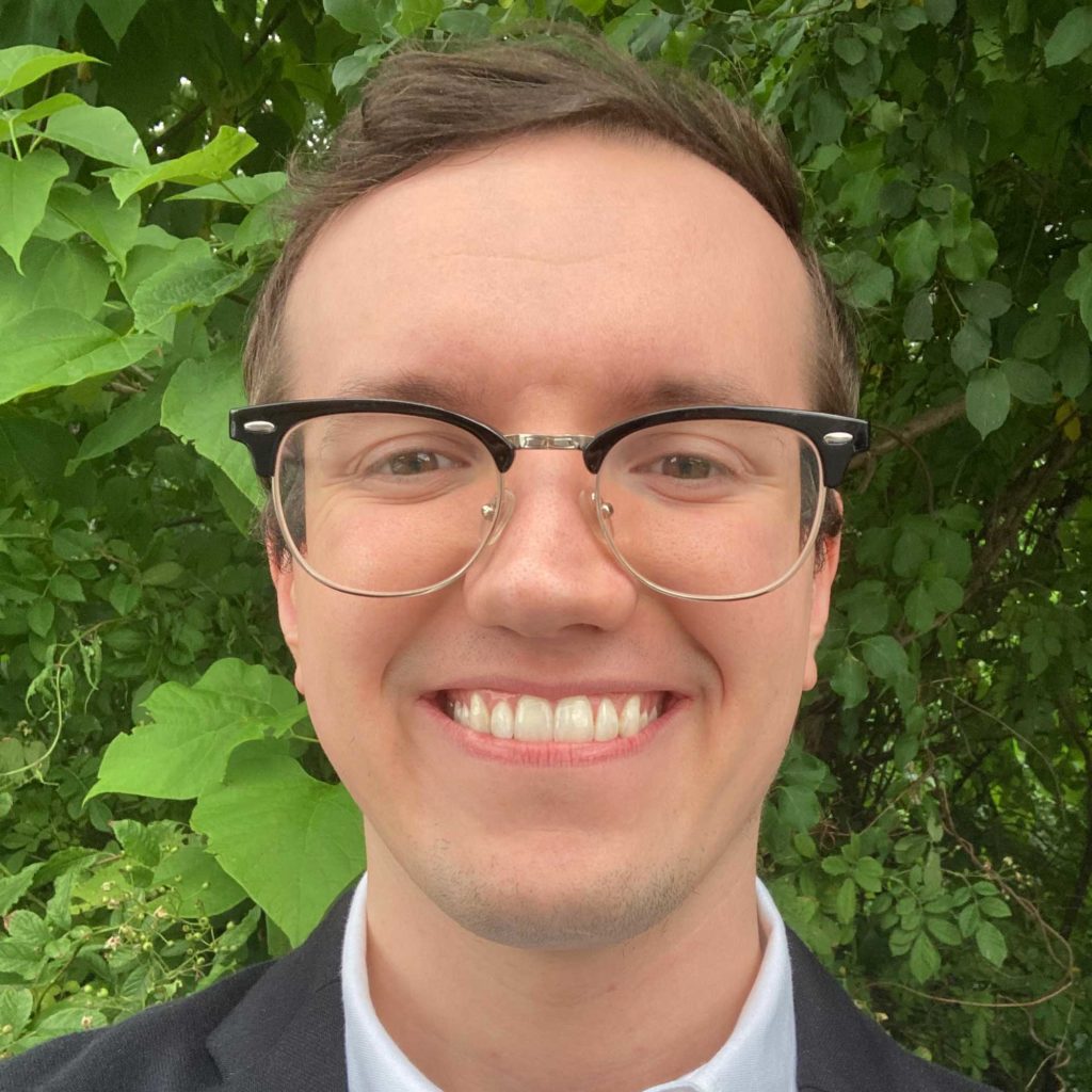 CDT's Aaron Spitler, wearing glasses and a dark suit jacket and white collared shirt, in front of a leafy green background.