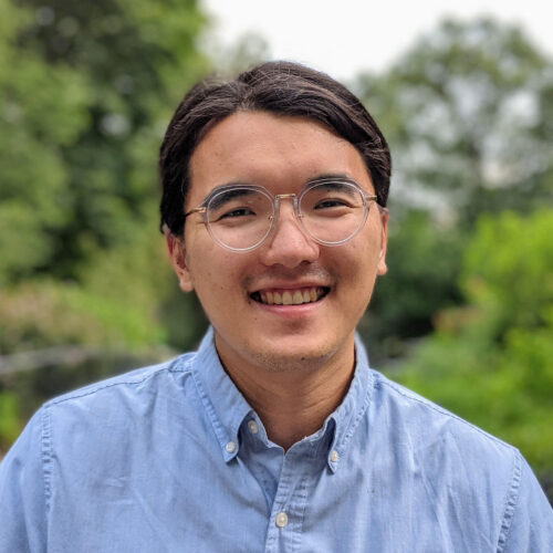 CDT Fellow Michael Yang, wearing a blue collared shirt and glasses, smiling outdoors in front of a lush growth of greenery.