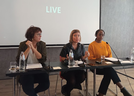 Photograph of three women seated at a table with microphones, speaking on a panel in front of a white screen that reads "Live." Asha Allen, at right of the image, wears glasses and an orange sweater. The woman in the middle wears a black short-sleeve button-up top, and the woman at left of the image wears a green collared jacket over a white blouse. 