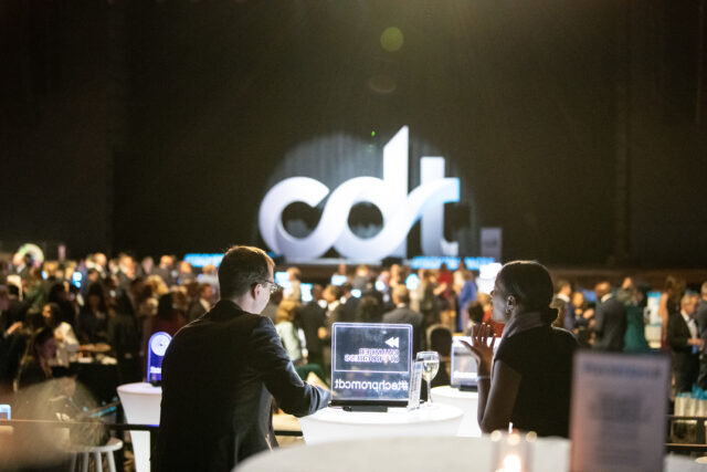 Two Tech Prom guests chat at a table, while looking out over the event. CDT logo spotlighted on stage.