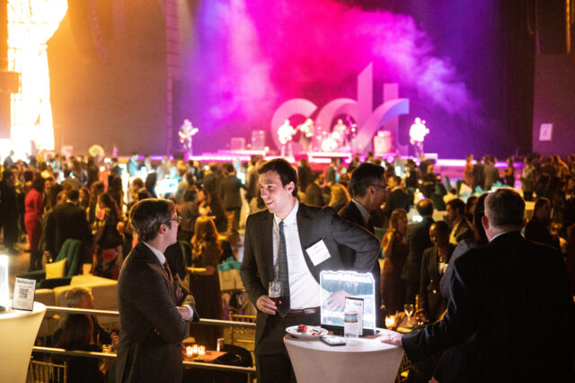 Two Tech Prom guests chat at a table, while looking out over the event. The stage is awash in music from the band and bright purple and red lights.