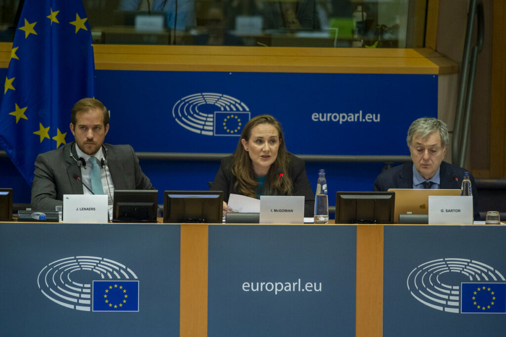 Photograph of two men and one woman seated on a table with microphones, speaking at a panel in front of a blue screen that reads ‘europarl.eu’, with the European Parliament sign and the EU flag. Iverna McGowan, seated in the middle, wears a black jacket and is speaking. The man on the left wears a grey jacket over a white shirt and a sky blue tie, and has earphones on his neck. The man on the right wears a dark blue jacket and a sky blue shirt, and sits in front of a laptop. Eric Vidal, 27/10/2022, © European Union 2022 - Source : European Parliament.