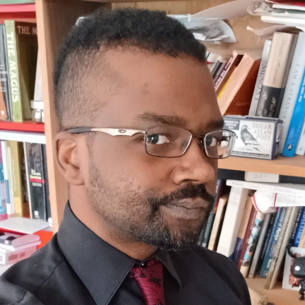 Damien Patrick Williams, wearing silver glasses with a black collared shirt and deep red tie. Standing in front of a bookcase.
