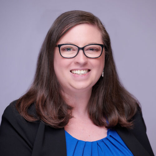 Elizabeth Dickson, smiling in dark rimmed glasses and wearing a blue top and black jacket, in front of a light purple background.