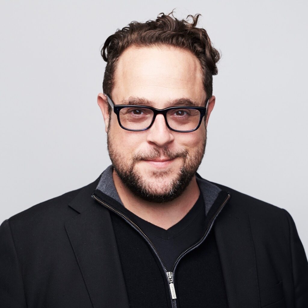 Kevin Bankston, wearing black glasses, a jacket and shirt in front of a white background.