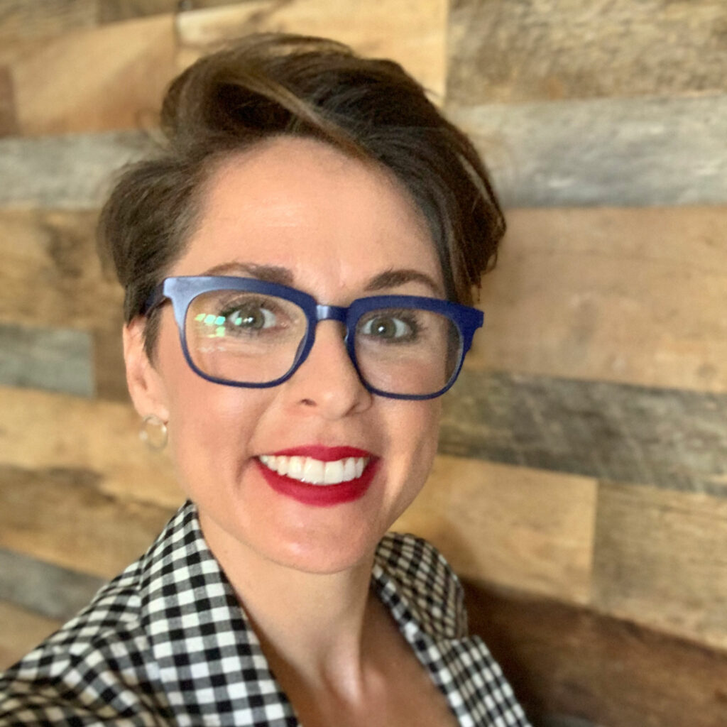 Amy Winecoff, smiling while wearing a checkered jacket, dark rimmed glasses and red lipstick in front of a wooden-paneled wall.