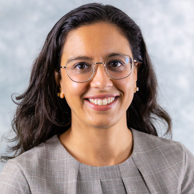 Priya Kumar, smiling wearing silver glasses and a grey top in front of a light colored background.
