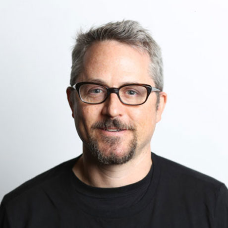 Sam Gregory, wearing black rimmed glasses and a black t-shirt, in front of a white wall.