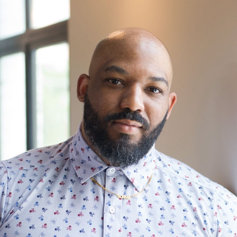 AJ is a bald Black male with a beard leaning against a wall by a window. He is wearing a short sleeve light blue button-down shirt with small red, white, and dark blue skulls patterned all over the shirt.