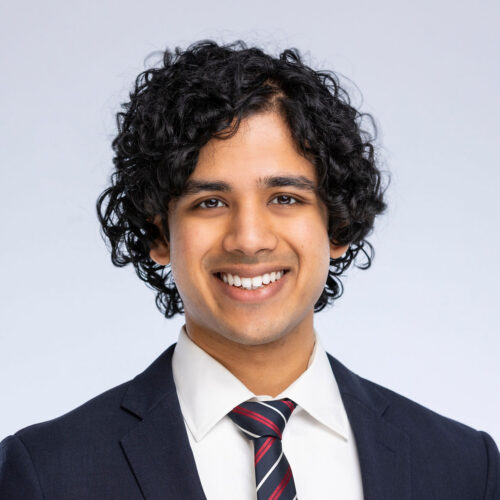 Chinmay Deshpande, smiling wearing a dark suit jacket and red, white and blue striped tie.