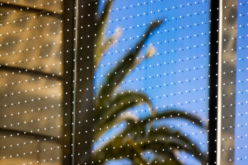 Reflective glass surface with a grid of small white dots, reflecting a blurred image of a palm tree and a blue sky.