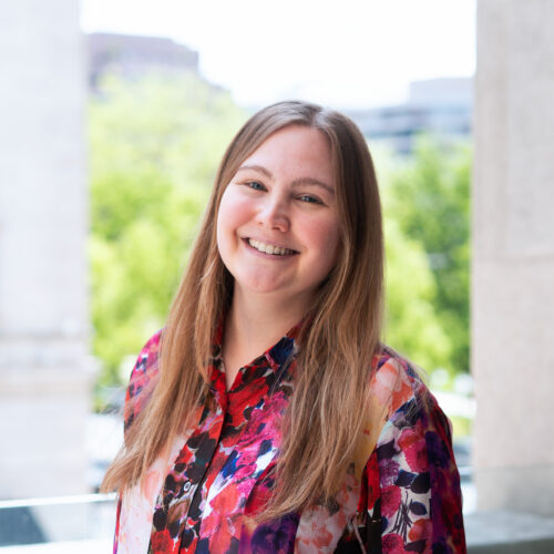 Elizabeth Seeger, smiling in a multi-color top, standing outside in a cityscape with trees behind her.