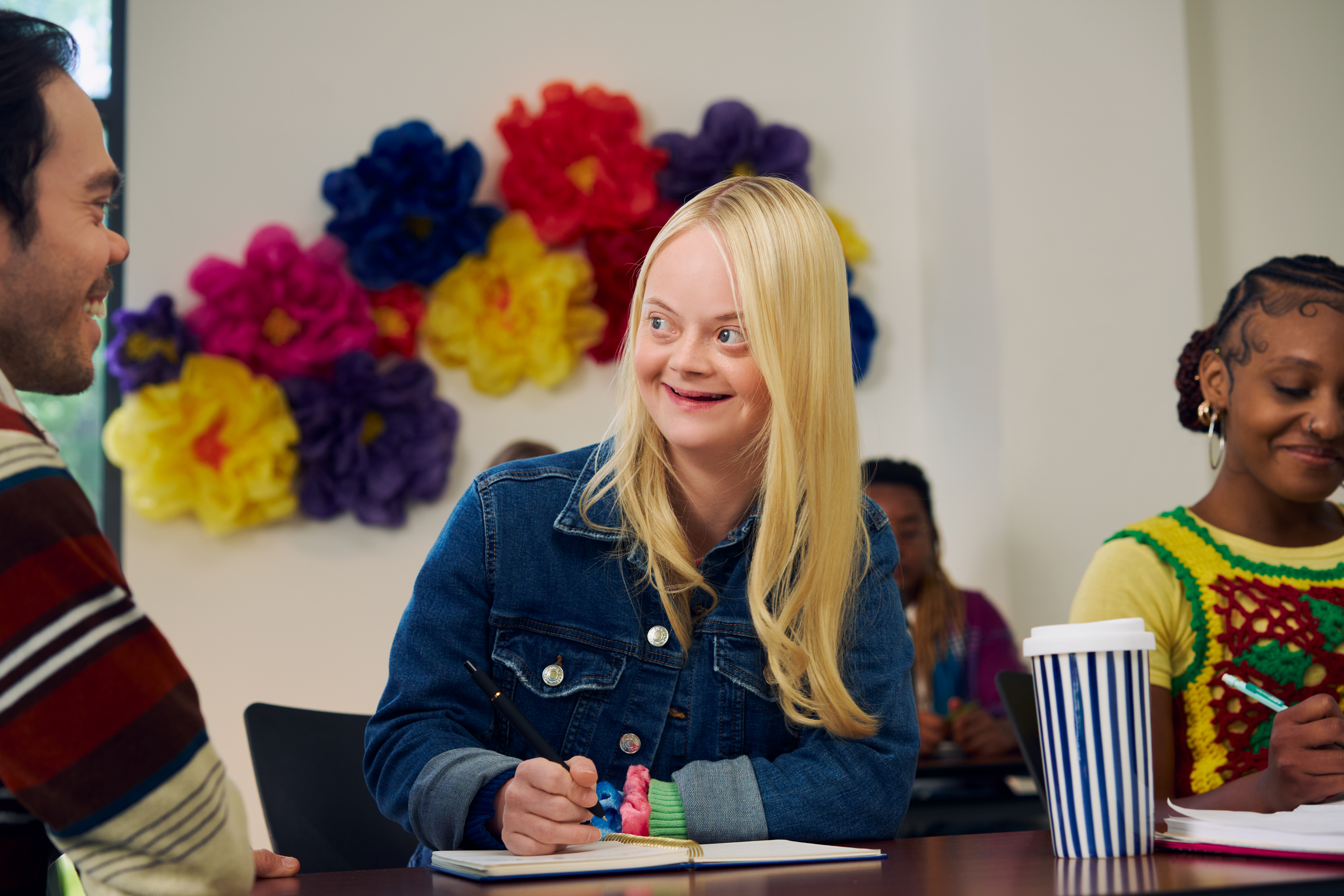 A Caucasian student with Down Syndrome sits at a desk happily talks with her professor, who has dwarfism, during class.