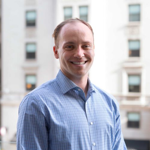 Quinn Anex-Ries, smiling outside in front of a city background. Wearing a lightly striped blue collared shirt.