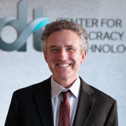 Travis Hall, smiling in a black jacket, white collared shirt and red tie in front of a CDT logo.