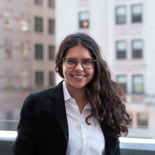 Ariana Aboulafia, smiling outside in front of a city background. With long dark hair, wearing a white collared shirt and black jacket.