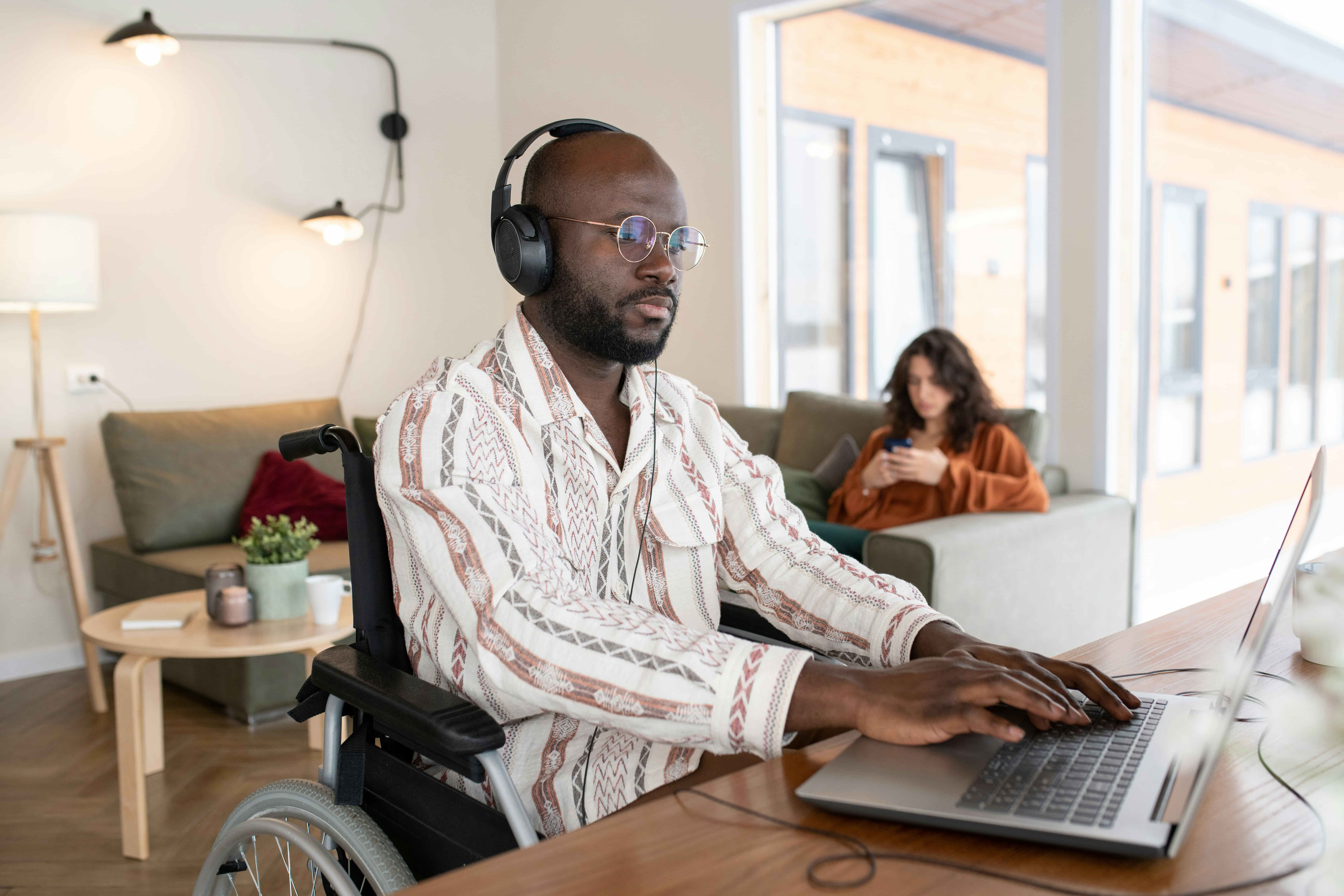 Black man with glasses and wearing a patterned shirt, in a wheelchair and using a laptop and headphones in a room with others.