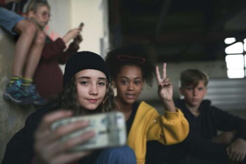 A group of young people, dressed in various hats and hoodies, sitting together indoors and talking while taking a selfie together.