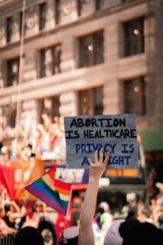 A photo of a group of people holding signs and waving flags, including a Pride flag and a sign saying “Abortion is Healthcare, Privacy is a Right.”