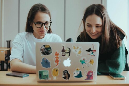 Two students in a class at university, sitting at a table with a laptop, covered in bright stickers.