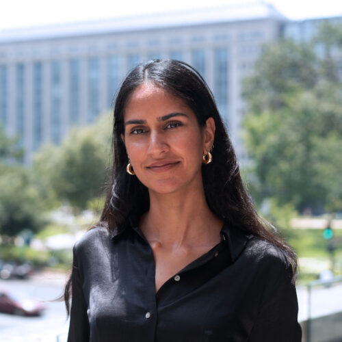 Aliya Bhatia, smiling outdoors wearing a black silk top and gold earrings.