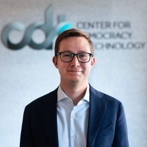 Matt O’Shaughnessy, in glasses and a dark suit jacket and white lightly striped dress shirt, in front of the CDT logo.