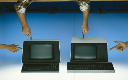 A pair of hands pointing and gesturing at old computer terminals and keyboards, on a blue background.