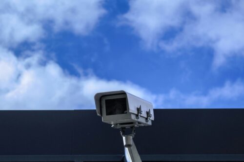 A white security camera mounted on a wall, active at daytime.