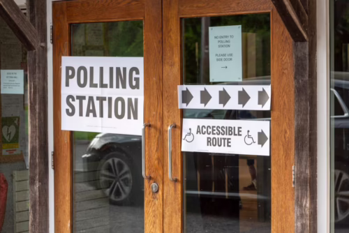 Double wooden doors at a polling station with printed signs directing voters to use a side entrance or the accessible route.